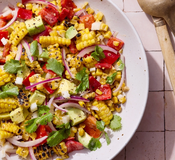 A vibrant salad with grilled corn, avocado, red onion, tomatoes, and cilantro on a white plate, set on a tiled surface with a spoon nearby.