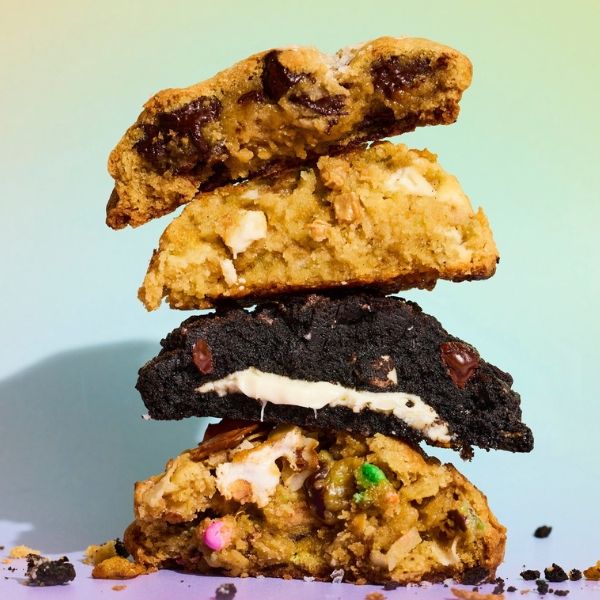 A stack of four assorted cookies with chocolate chips and colorful candies, set against a pastel gradient background.