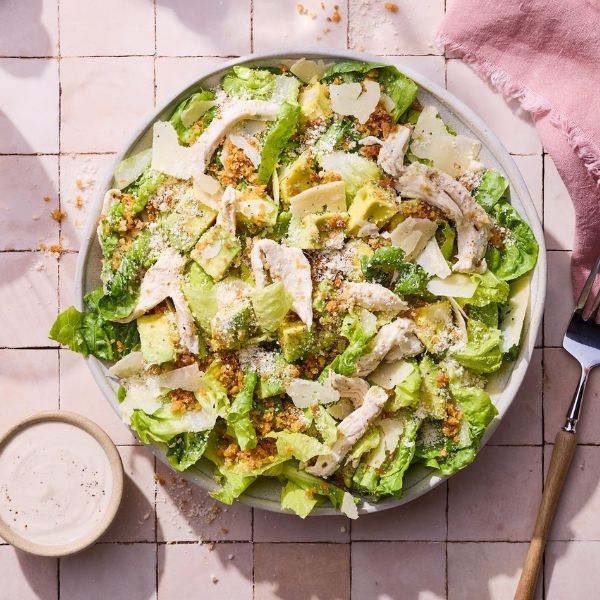 A Caesar salad with grilled chicken, romaine lettuce, croutons, Parmesan cheese, and dressing on a tiled surface with a pink napkin.