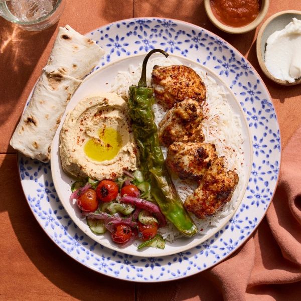 A plate with grilled chicken, rice, hummus, grilled pepper, tomato salad, flatbread, and sauces on a patterned dish.