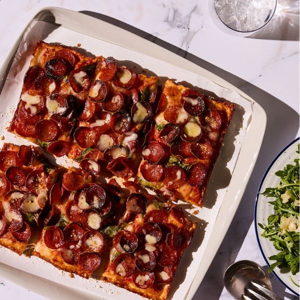 Square pepperoni pizza with melted cheese on a white tray, next to a plate of arugula salad with parmesan and a glass of water.