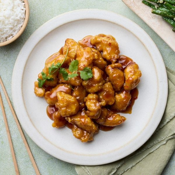 Plate of glazed orange chicken garnished with cilantro, served with a side of rice and chopsticks on a green napkin.