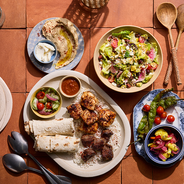 A table with grilled meat, rice, salad, hummus, yogurt, flatbreads, and pickled vegetables, set on a terracotta surface with spoons and wooden utensils.