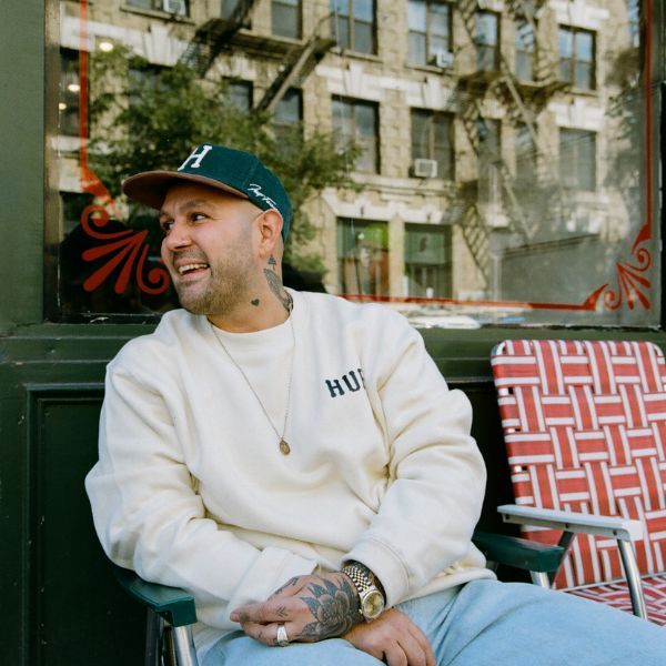 Frank Pinello in casual clothing and hat, smiling, sitting on a red patterned chair outside a building with large windows.