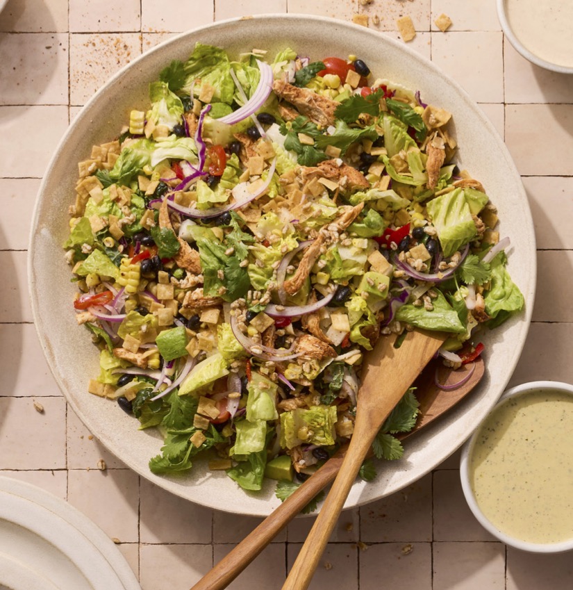 A vibrant salad with lettuce, shredded chicken, red onions, corn, black beans, and tortilla strips in a bowl, with dressing on the side.