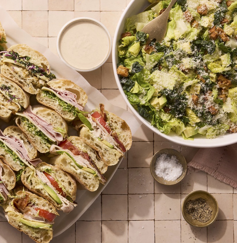 A platter of assorted sandwiches with lettuce and a bowl of Caesar salad, accompanied by a small dish of salt and pepper on a tiled surface.