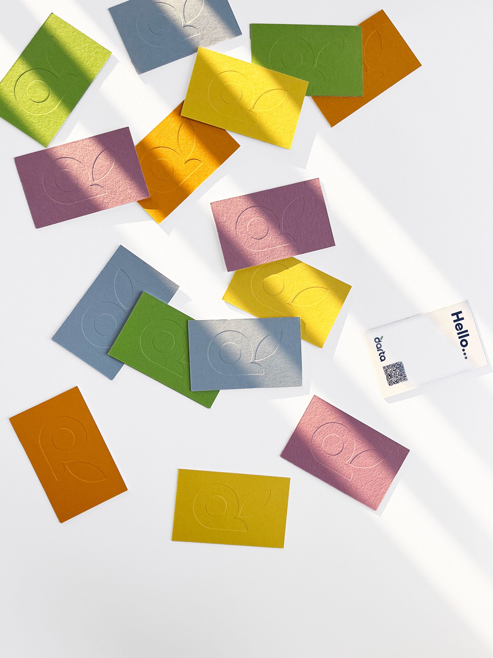 Scattered colorful embossed cards on a white surface with sunlight, including green, blue, yellow, and purple hues. A "Hello" card with a QR code.