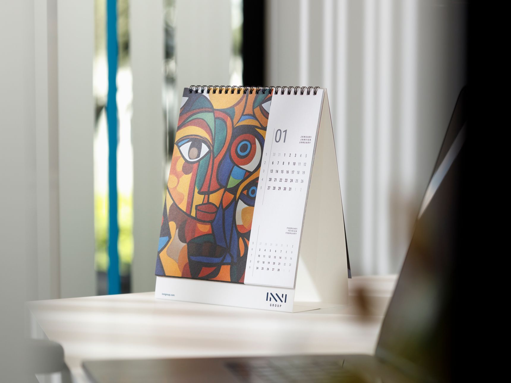 A desk calendar with abstract art on the cover is displayed on a white table next to a laptop, with blurred background elements.