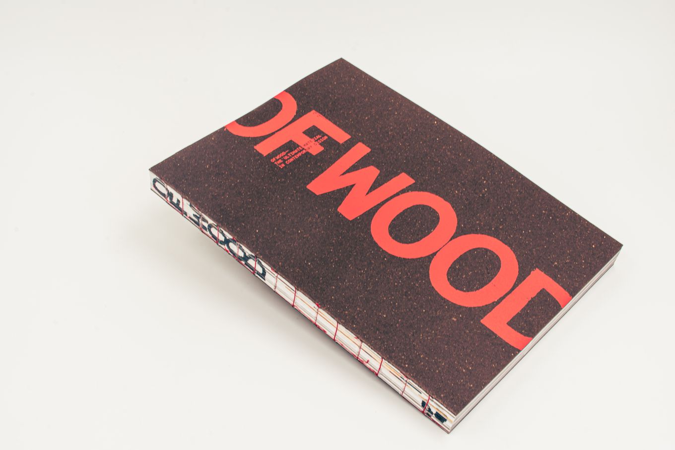 A book with a dark brown cover titled "OF WOOD" in large red letters, resting on a light surface.