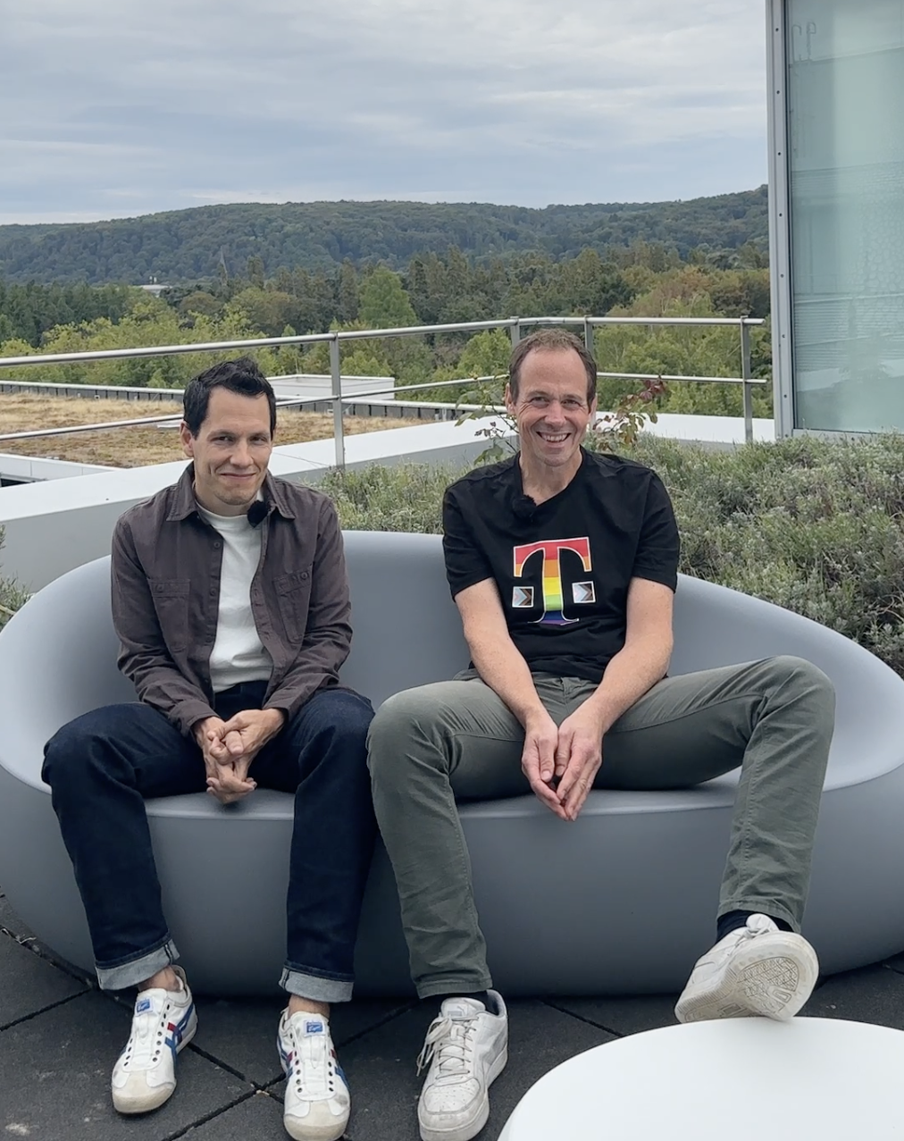 Two men sit on a gray outdoor sofa on a rooftop terrace, with a scenic view of trees and hills in the background.
