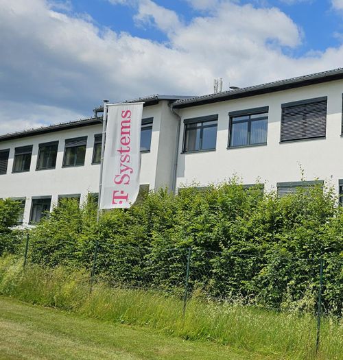 White building with a "T-Systems" banner, surrounded by greenery under a partly cloudy sky.