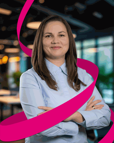 Woman in a blue shirt smiling, arms crossed, with a pink ribbon spiraling around her, standing in a modern office setting.