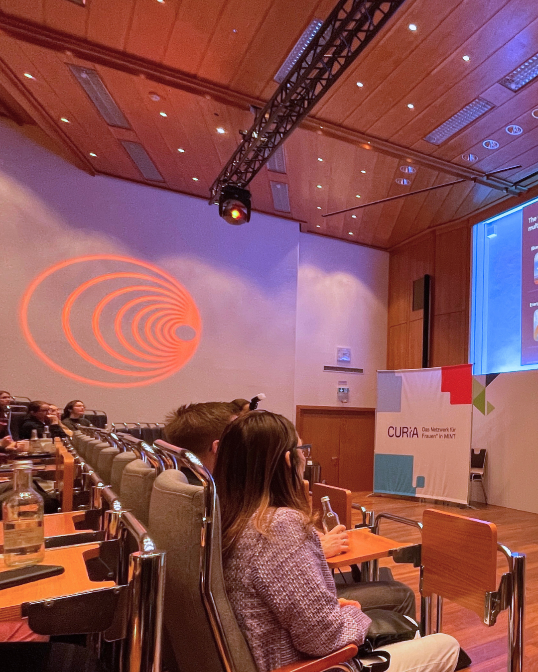 Audience seated in a modern conference room with wooden ceiling, projected presentation slides, and red spotlight design on the wall.