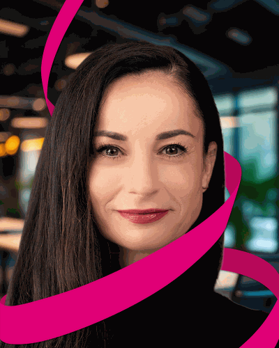 Woman with long dark hair and red lipstick, wrapped in a pink ribbon, smiling in a modern office setting with blurred background.