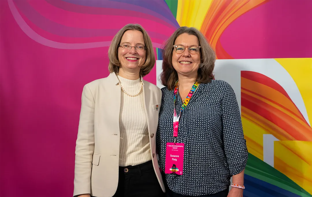 Two women smiling in front of a vibrant, colorful magenta background. One wears a white blazer, the other a patterned shirt with a magenta lanyard.