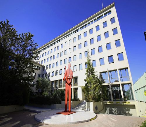 Modern white building with many windows, featuring a tall red statue of a person in front, surrounded by trees under a clear blue sky.