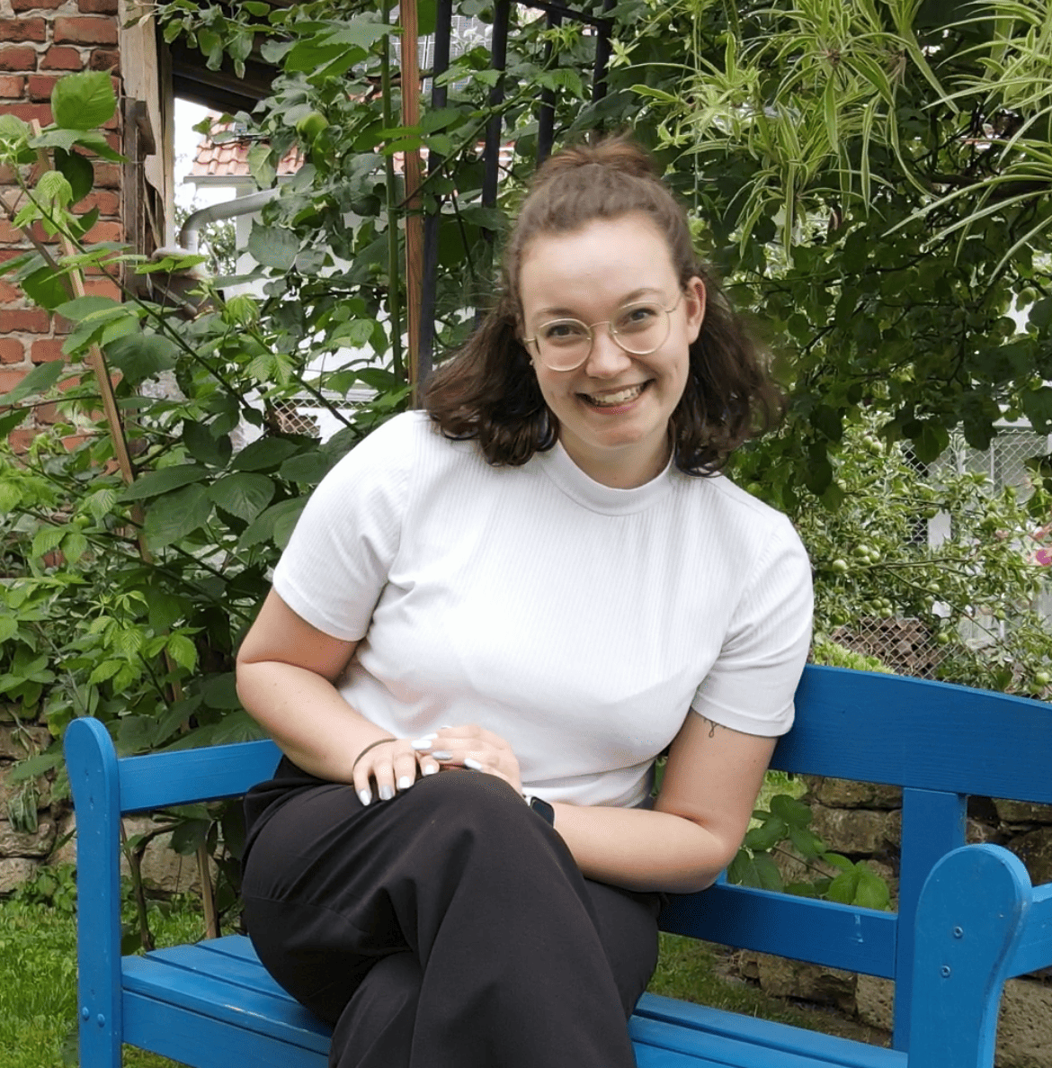 A young woman with glasses, wearing a white shirt, black wide-leg pants, and white sneakers, is sitting and smiling on a bright blue bench in a garden surrounded by greenery