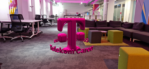 Modern office space with pink lighting, featuring desks, chairs, a gray sectional sofa, colorful ottomans, and a large Telekom Career logo.