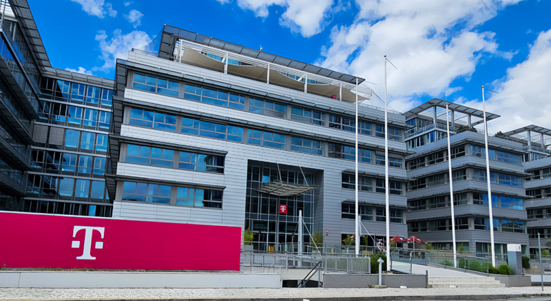 Modern office building with glass windows and a large magenta T-Mobile logo in front, under a partly cloudy sky.
