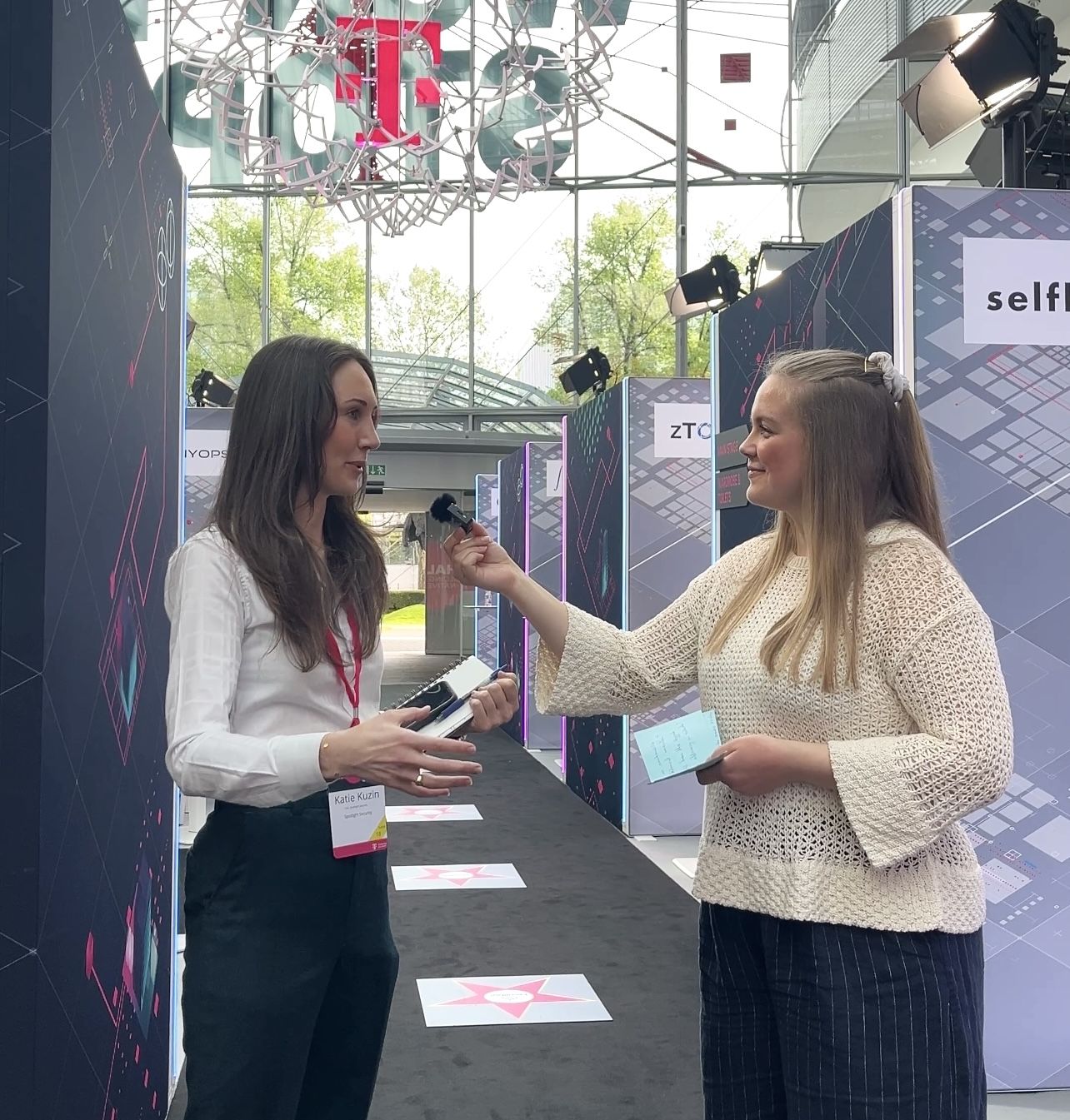 Two women in conversation at an event, one holding a microphone. Brightly lit space with geometric displays in the background.