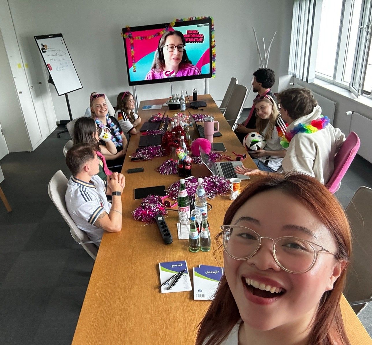 Group of people in a decorated conference room video chatting with someone on screen; one person taking a selfie, others smiling and chatting.