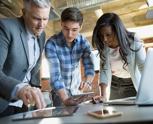 Three colleagues collaborate around a table with digital devices, focused on a tablet. They are in a modern office setting.