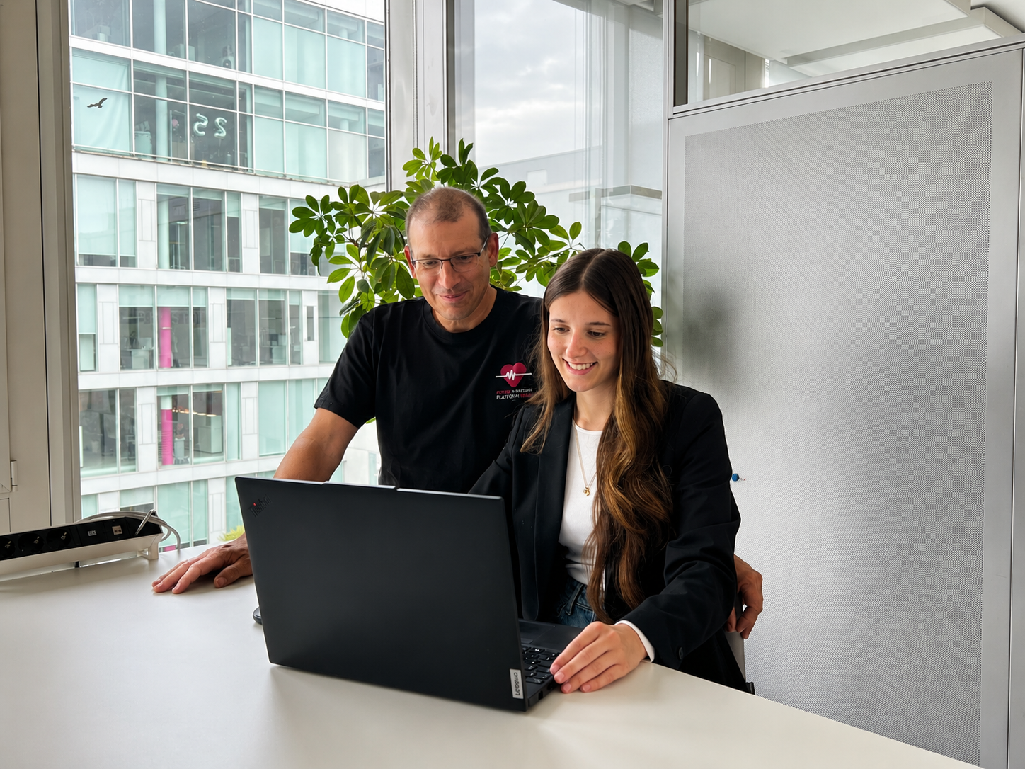 Two people smiling while looking at a laptop in an office with a large window and a potted plant nearby.