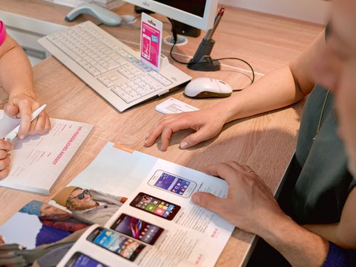 Two people at a desk discussing a brochure featuring smartphones. A computer keyboard and mouse are visible in the background.