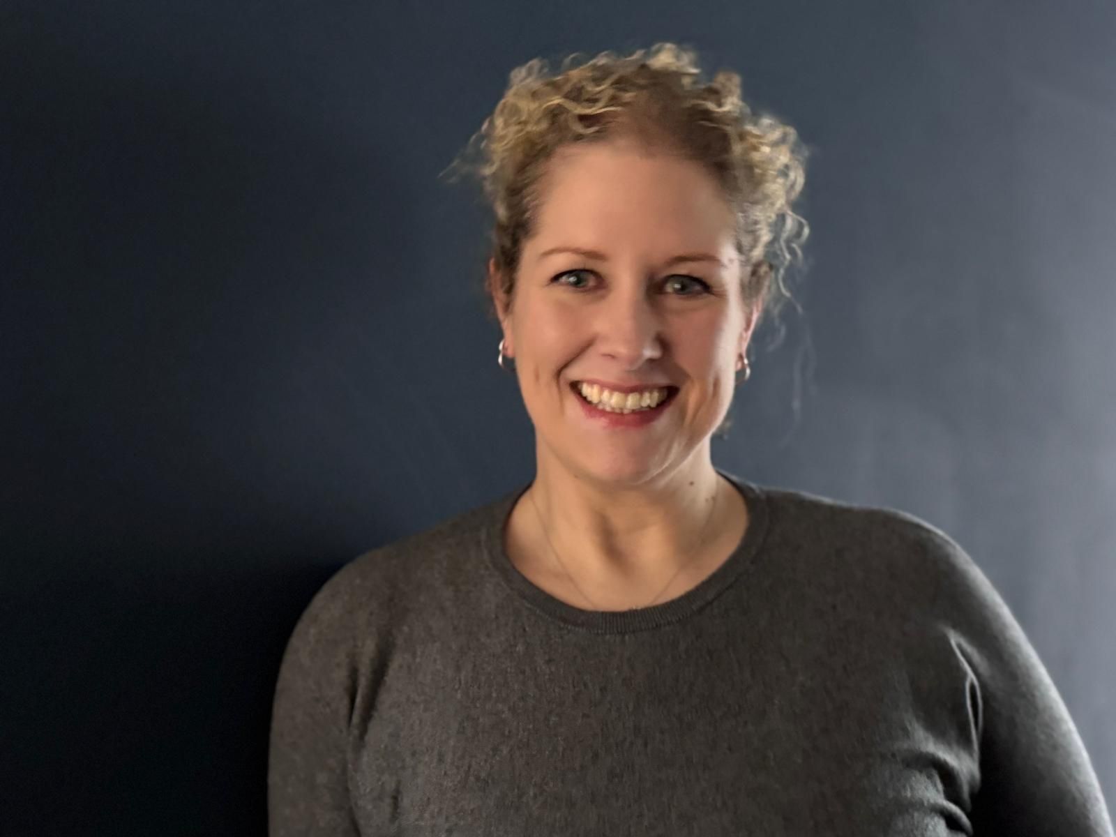 Smiling person with curly hair wearing a gray sweater, standing against a dark background.