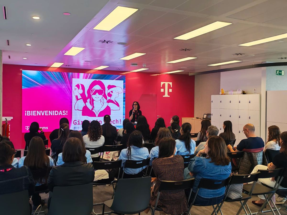 A group of people sit in a conference room facing a speaker and a large screen displaying "¡Bienvenidas!" and "Girls in Tech!" in a T-Mobile setting.
