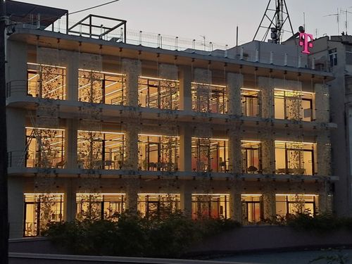 A building with large windows illuminated from inside at dusk, displaying a vertical garden facade. A magenta "T" logo is visible on the roof.