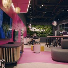 Modern lounge with pink accents, greenery wall, seating, and people conversing. "Ahoj Bratislava" text on the wall.