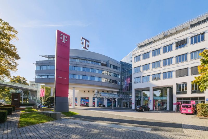 Modern office buildings with the Deutsche Telekom logo, featuring a curved glass facade and a magenta sign, under a clear blue sky.