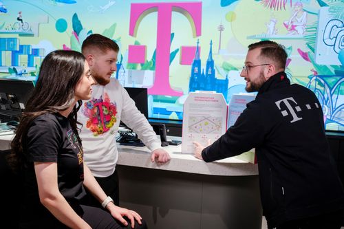 Three people in a store discussing a brochure, with a large magenta "T" logo and colorful background behind them.