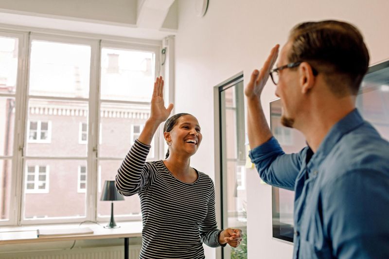Two people in a bright room giving a high five, smiling at each other. One wears a striped shirt, the other a blue shirt and glasses.