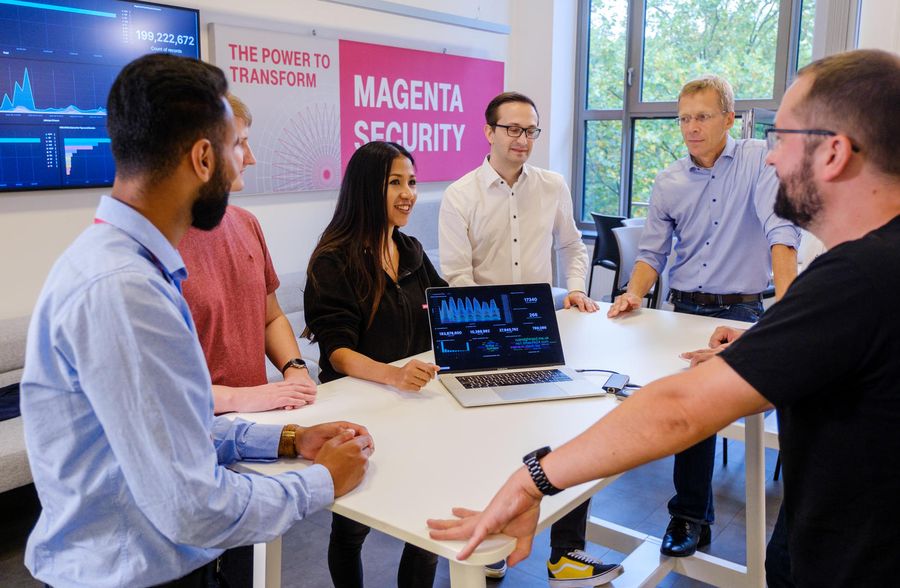 A group of five people having a discussion around a table with a laptop, in a modern office setting with large windows.