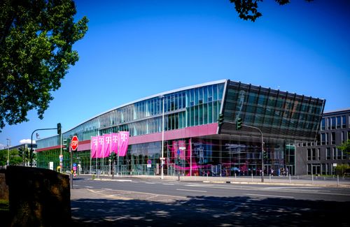 Modern glass building with magenta banners under a clear blue sky, surrounded by trees and a street with traffic lights.