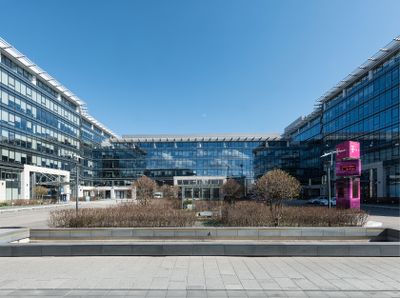 Modern office building with glass facade, featuring a central courtyard and a magenta sign with logo. Clear blue sky in the background.