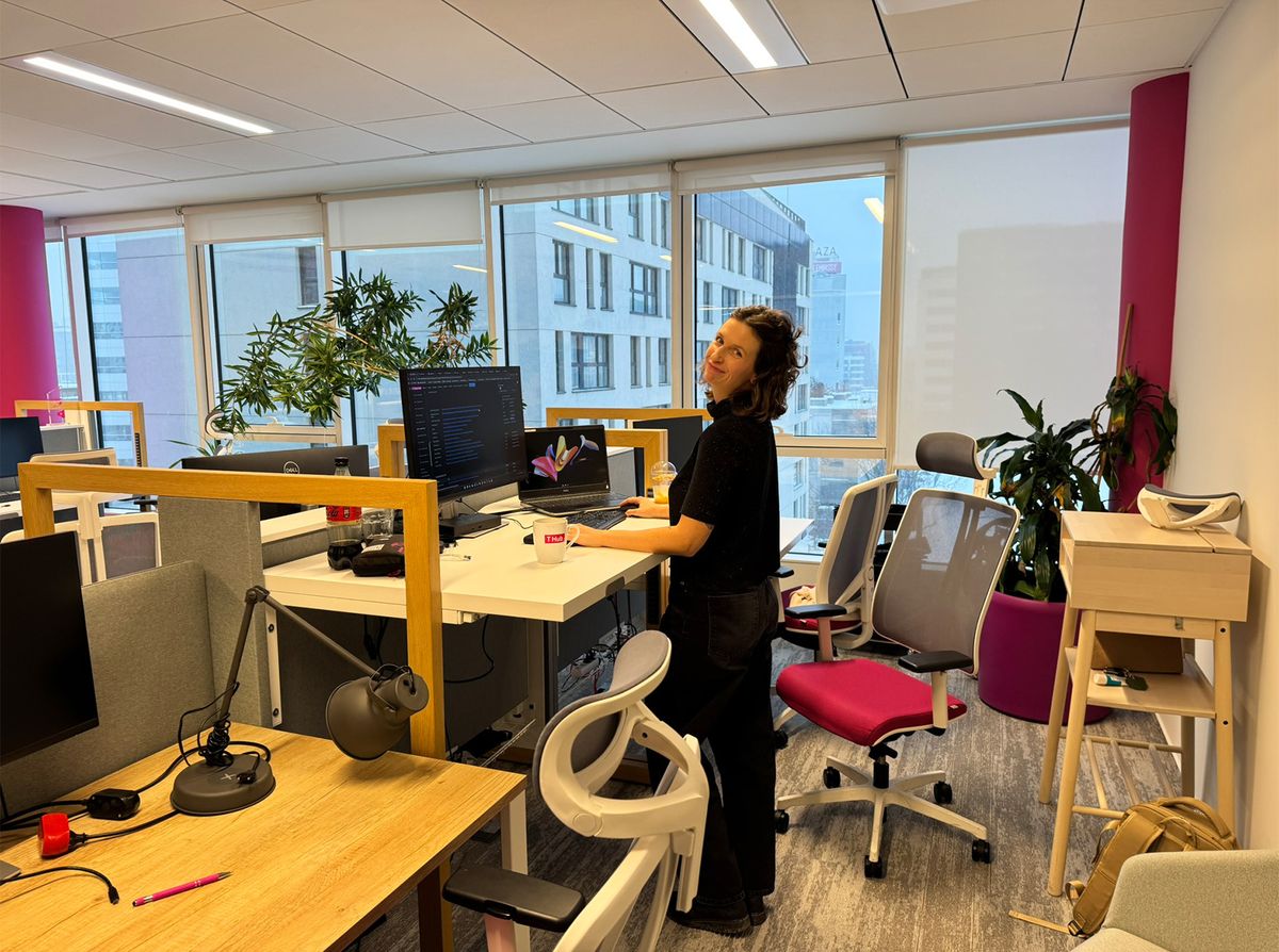Woman standing at a desk in a modern office with large windows, multiple monitors, and potted plants nearby.