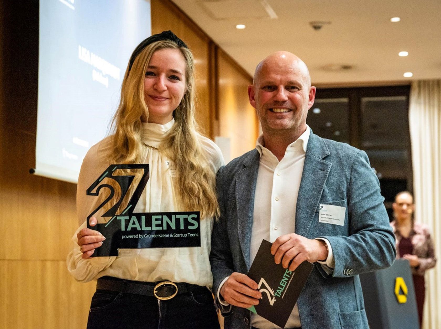 Two people smiling and holding "27 Talents" awards, standing indoors at an event with a presentation screen behind them.