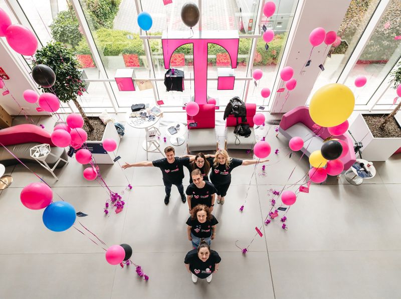Group of people in black shirts stand among colorful balloons in a modern, glass-walled room, with a large magenta logo.