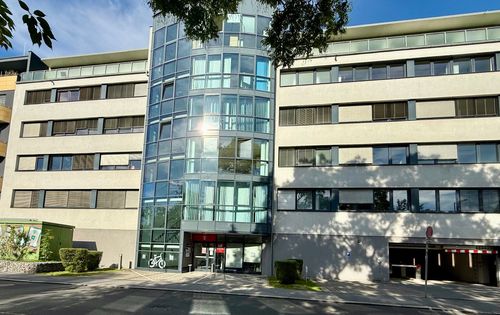 Modern building with a central glass facade, surrounded by trees and a clear blue sky. Sunlight reflects off the windows.