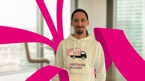Man with long hair in a white hoodie featuring a delivery truck graphic. Magenta abstract shapes surround him; blurred office setting in background.