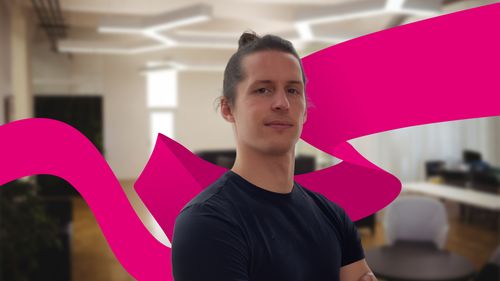 Young man with long hair in a bun, wearing a navy shirt, standing  in a modern office with a magenta wave design.