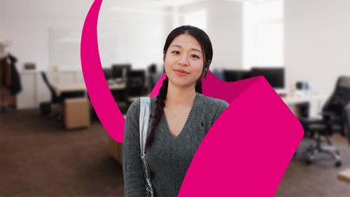 A woman with braided hair stands smiling in a modern office setting, with a magenta abstract shape in the foreground.