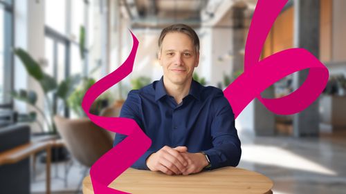 Smiling man in a blue shirt sits at a table in a modern office with large windows and plants, with magenta ribbon-like graphics around him.