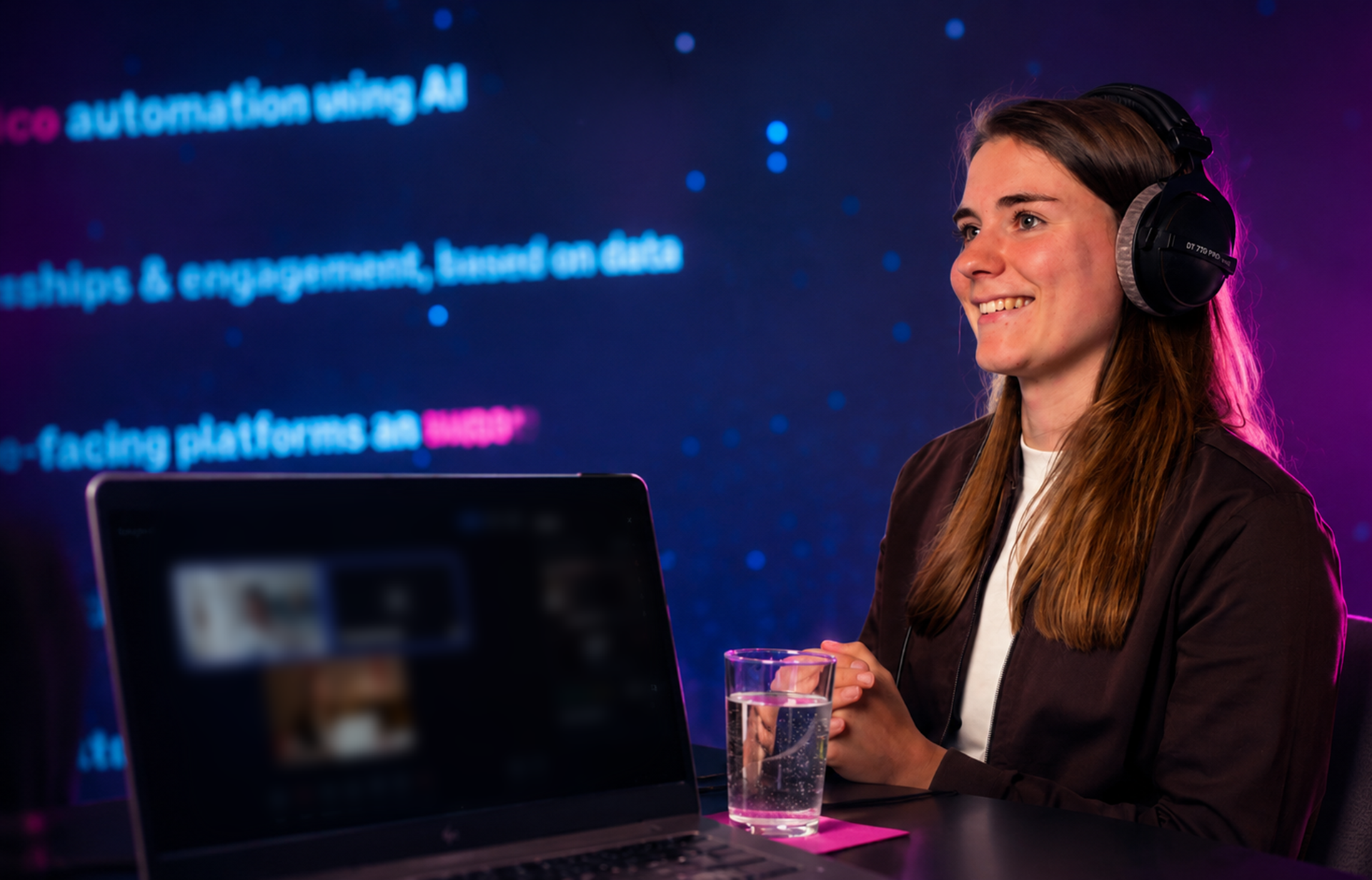 A woman wearing headphones sits at a desk with a laptop and a glass of water, smiling against a backdrop of blue and pink text.