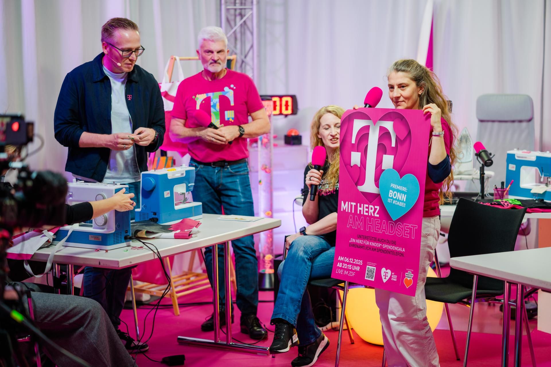 Group at a pink charity broadcast: woman holds a large heart-shaped "T" poster while others use microphones and sewing machines.