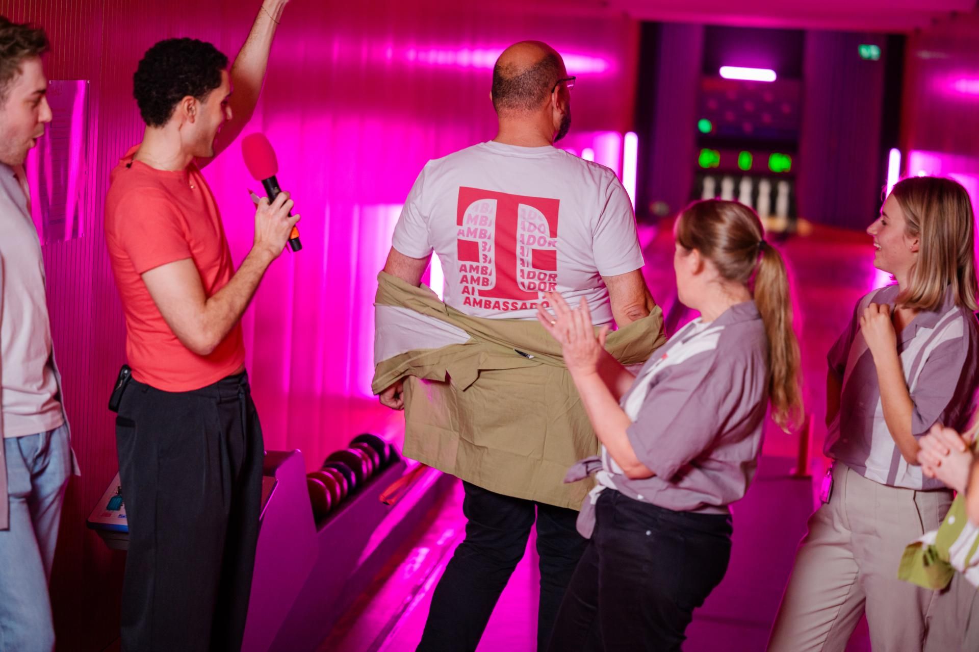 People cheering in pink-lit bowling alley as a man reveals an "Ambassador" t-shirt on his back while another holds a microphone.
