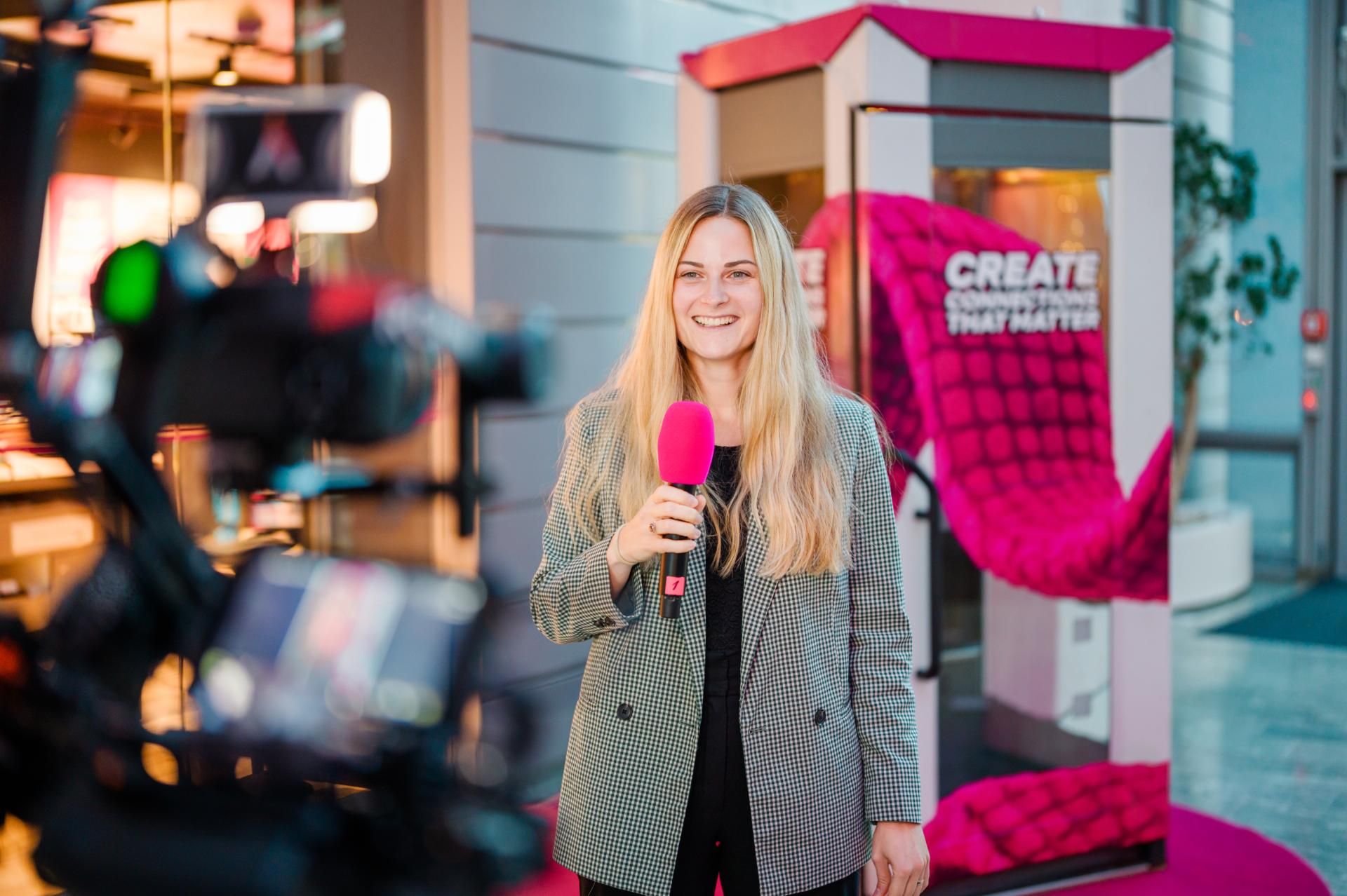 Smiling blonde woman holding a bright pink microphone while being filmed in front of a pink promotional display.
